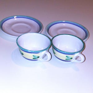 May include: Two white ceramic teacups with blue and green trim and a yellow band. The cups have a floral design and are sitting on matching saucers.