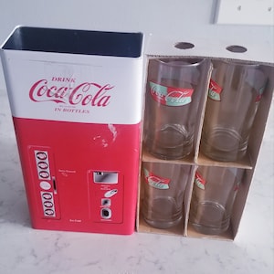 Puede incluir: Réplica de máquina expendedora de Coca-Cola roja y blanca con un dispensador de vidrio y una caja de cuatro vasos de Coca-Cola. Los vasos tienen un logotipo de Coca-Cola rojo y verde.