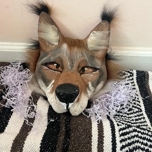 May include: A handmade fox mask with brown, gray, and white fur details. The mask has pointed ears with black tufts, and is resting on a black, white, and brown patterned blanket. The eyes are a golden brown color.