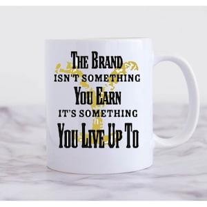 May include: White ceramic mug with a black and gold design. The text on the mug reads "The brand isn't something you earn, it's something you live up to."