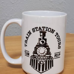 May include: White ceramic mug with a black and white graphic of a train engine with the text "Train Station Tours" around the engine and "Team" and "RIP" on either side of the engine.
