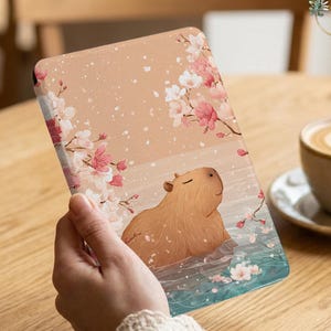 May include: A tablet case featuring a serene illustration of a capybara in water, surrounded by cherry blossoms. The design has a soft, pastel color palette with pink, white, and blue accents. The case is held by a person.