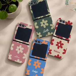 May include: Four foldable phone cases featuring floral patterns and personalised names. The cases are dark green with cream flowers, dusty pink with cream flowers, blue with orange flowers, and cream with red flowers. Each case has a black screen area.