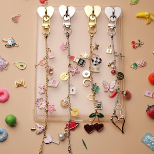 May include: Four keychains with heart-shaped clasps and decorative charms. Charms include a lipstick, a star, a lock, a butterfly, and a heart. The background is a light tan color with scattered miniature food and object charms.