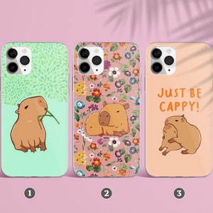 Puede incluir: Tres fundas para teléfono con diferentes diseños que presentan capibaras. La primera funda tiene un fondo verde con una capibara comiendo una hoja. La segunda funda tiene un fondo floral con una capibara y una capibara más pequeña. La tercera funda tiene un fondo rosa con dos capibaras abrazándose y el texto "Just be Cappy!"