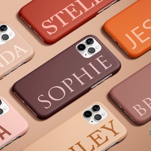 Könnte beinhalten: Sortiment an personalisierten Handyhüllen mit verschiedenen Namen, die in Weiß, Rosa und Orange auf verschiedenen farbigen Hintergründen gedruckt sind. Die Hüllen sind auf einer beigefarbenen Oberfläche angeordnet.