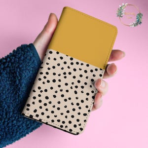 May include: A mustard yellow and cream colored notebook with a black polka dot pattern. The notebook is closed and held in a hand.