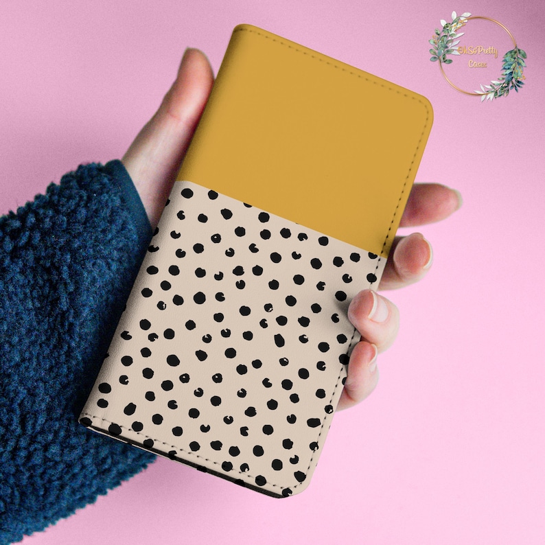May include: A mustard yellow and cream colored notebook with a black polka dot pattern. The notebook is closed and held in a hand.