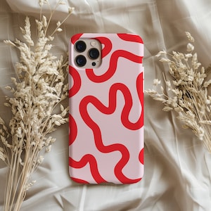 Pode incluir: Uma capa de telefone rosa com um padrão ondulado vermelho. A capa tem um acabamento brilhante e um recorte para a câmera. O design é de estilo moderno e abstrato. A capa é rodeada por flores secas.