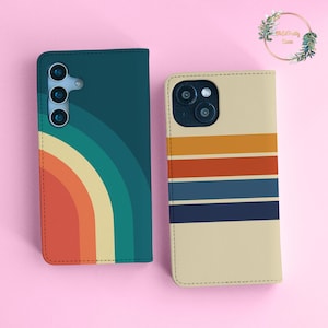 May include: Two rectangular phone cases. The left case is dark teal with a curved, multi-colored rainbow pattern in orange, yellow, and teal. The right case is cream-colored with horizontal stripes in orange, red, blue, and dark navy blue.