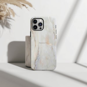 Puede incluir: Funda de teléfono con estampado de mármol blanco y beige con el nombre "MEREDITH" impreso en fuente negra fina. La funda tiene un marco de lente de cámara negro y botones laterales negros. La funda se muestra sobre una superficie blanca.
