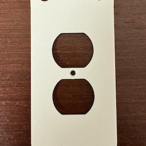 Może przedstawiać: Biała dekoracyjna osłona gniazdka elektrycznego z falistym brzegiem i jednym otworem na gniazdko.