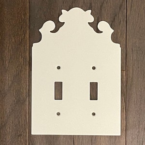 Könnte beinhalten: Weiße Keramik-Wandplatte mit einem dekorativen Oberteil und zwei Ausschnitten für Lichtschalter.
