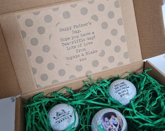 May include: A brown cardboard box with green shredded paper inside. The box contains three golf balls. One golf ball has the text "World's Best Dad" printed on it. Another golf ball has a photo of a man and a girl printed on it. The box also includes a card with the text "Happy Father's Day. Hope you have a Tea-riffic day! Lots of love from Sophie & Blake XXX".