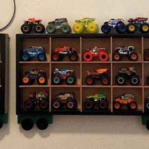 Może przedstawiać: Czarna drewniana p&oacute;łka z wieloma przegr&oacute;dkami zawiera kolekcję zabawek monster truck&oacute;w. Ciężar&oacute;wki są w r&oacute;żnych kolorach, w tym czerwonym, niebieskim, zielonym, ż&oacute;łtym i czarnym. P&oacute;łka jest zamontowana na ścianie.