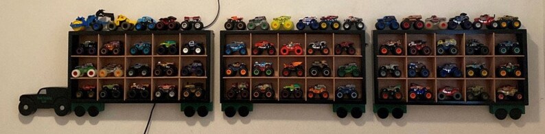 Może przedstawiać: Czarna drewniana p&oacute;łka z wieloma przegr&oacute;dkami zawiera kolekcję zabawek monster truck&oacute;w. Ciężar&oacute;wki są w r&oacute;żnych kolorach, w tym czerwonym, niebieskim, zielonym, ż&oacute;łtym i czarnym. P&oacute;łka jest zamontowana na ścianie.