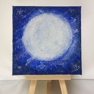 May include: Square canvas art depicting a white, textured full moon against a dark blue, starry background. The painting is displayed on a small wooden easel, creating a celestial theme.