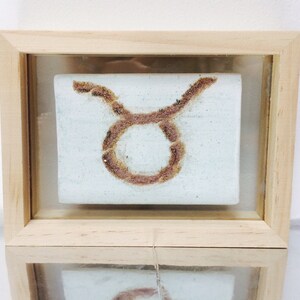 May include: A framed artwork featuring the Taurus zodiac symbol. The symbol is a brown, textured design on a light blue background. The frame is made of light-colored wood, and the artwork is displayed behind glass.