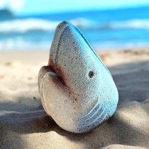 Puede incluir: Una figurita de tiburón de cerámica azul y blanca con una superficie texturizada. La figurita está sentada en una playa de arena.