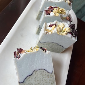 May include: Four rectangular bars of handmade soap are arranged on a white tray. The soap has a two-tone design, with a light blue top and a speckled green base, separated by a dark line. Dried flowers and petals adorn the top of each bar.