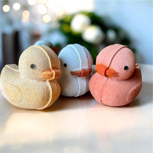 Puede incluir: Tres bombas de baño con forma de pato. La primera es de color amarillo claro, la segunda es blanca y la tercera es rosa. Cada una tiene un pico naranja y ojos negros. Las bombas de baño están sobre una superficie blanca.