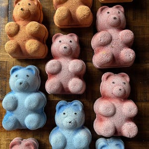 May include: A selection of bear-shaped bath bombs in shades of orange, pink, and blue, arranged on a wooden surface. The bath bombs are designed with bear-like features, offering a colourful display.