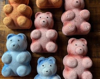 Könnte beinhalten: Draufsicht auf mehrere bärenförmige Badebomben in verschiedenen Farben, darunter Orange, Rosa und Blau. Die Badebomben sind auf einer Holzoberfläche angeordnet und zeigen ihre detaillierten Bärenmerkmale.