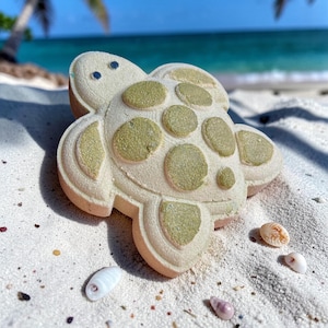 Könnte beinhalten: Eine schildkrötenförmige Badesalzkugel mit hellbeigem Körper und olivgrünen Flecken, auf weißem Sand mit Muscheln. Im Hintergrund eine tropische Strandlandschaft mit Palmen und türkisfarbenem Wasser. Ca. 10 cm lang.
