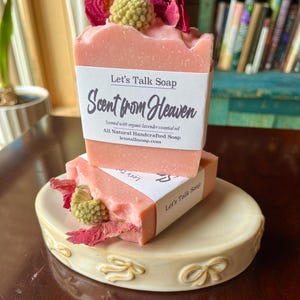 Puede incluir: Dos barras de jabón rosa con el texto "Let's Talk Soap" y "Scent from Heaven" en una etiqueta blanca. El jabón está decorado con flores secas y se encuentra en una jabonera color crema.