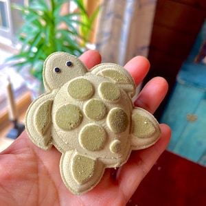 Puede incluir: Una bomba de baño con forma de tortuga verde claro con una superficie texturizada y dos pequeños puntos negros para los ojos. La bomba de baño tiene un diseño detallado con círculos en relieve en la espalda.