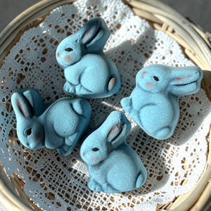 May include: Four light blue bunny figurines with dark brown ears and eyes, and pink cheeks, are arranged in a woven basket atop a white lace doily. The bunnies are in various poses, creating a charming Easter display.