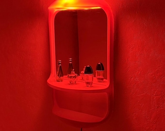 Puede incluir: Un espejo de tocador de pared de plástico rojo con un estante integrado y un borde superior redondeado. El espejo refleja varios frascos de perfume de vidrio pequeños colocados en el estante. Un cable de alimentación negro cuelga de la unidad.