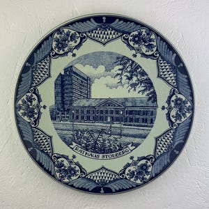 Op de afbeelding: Een ronde, blauw-witte decoratieve plaat. De plaat heeft een gedetailleerde illustratie van een gebouw en het omliggende landschap. De woorden "RATHAUS STOLBERG" staan op de plaat gedrukt. De plaat heeft een decoratieve rand.