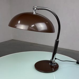 May include: A brown desk lamp with a curved, chrome-colored arm and a rounded lampshade. The lamp sits on a brown circular base. The lamp is on a white table with a light blue glass top.