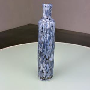 Op de afbeelding: Een hoge, slanke keramische vaas met een textuur die lijkt op houtnerf. De vaas is voornamelijk lichtblauw met donkerblauwe en bruine accenten. Hij staat op een lichtgroen oppervlak.