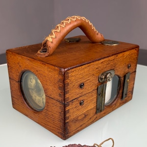May include: A wooden box with a leather handle and metal details. The box has a circular window on the side and a rectangular window on the front. A small maroon pouch and two black metal rods are in front of the box.
