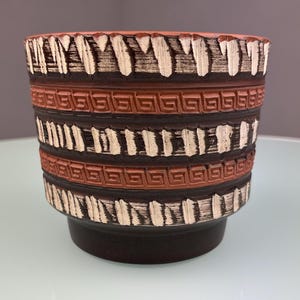 Op de afbeelding: Een bruine keramische plantenbak met een geometrisch ontwerp. De plantenbak heeft afwisselende banden van donkerbruine, lichtbruine en witte patronen. De bovenrand heeft een getextureerde, gekartelde rand. De plantenbak staat op een licht uitlopende basis.