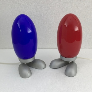 May include: Two table lamps with a unique design. The lamps are shaped like eggs with three legs. One lamp is blue and the other is red.