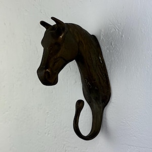 May include: A bronze-coloured horse head wall hook. The hook is shaped like a horse's head and neck, with a curved hook at the bottom. The horse head has detailed features, including the eyes, ears, and mane. The hook is mounted on a white wall.
