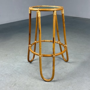 Op de afbeelding: Een vintage rotan barkruk met een ronde zitting en een stevige, open basis. De kruk heeft een natuurlijke, lichtbruine kleur met geweven details en een cirkelvormige voetensteun. De poten buigen sierlijk, wat bijdraagt aan de retro-uitstraling.