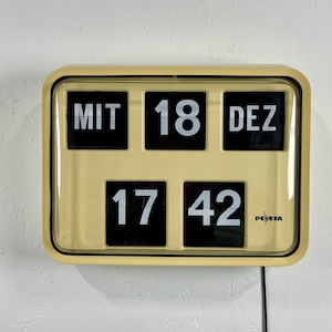 Puede incluir: Un reloj de números de volteo beige con números negros que muestran la fecha "MIT 18 DEZ" y la hora "17 42". El reloj tiene un cable negro y la marca "DEHESA" es visible.