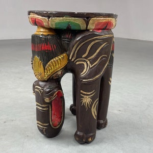 Op de afbeelding: Een handgesneden houten olifanten kruk met een ronde bovenkant. De olifant is geschilderd in tinten van bruin, rood, goud en groen. De kruk is een decoratief meubelstuk dat kan worden gebruikt als bijzettafel of voetenbank.