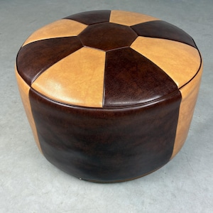 Op de afbeelding: Een ronde, tweekleurige poef in bruin en beige. De bovenkant heeft een geometrisch patroon met afwisselende bruine en beige secties. De zijkanten zijn voornamelijk bruin. Dit meubelstuk is ontworpen om op te zitten of als voetensteun.