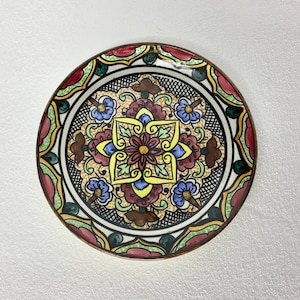 Op de afbeelding: Een klein, rond keramiek bord met een kleurrijk bloemenontwerp. Het bord heeft een zwarte achtergrond met een centraal stervormig patroon in tinten geel, rood en blauw. De rand van het bord is versierd met groene, rode en witte bloemenmotieven.