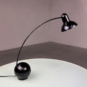 Op de afbeelding: Een zwarte bureaulamp met een gebogen arm en een ronde voet. De lamp heeft een kleine, koepelvormige kap en staat op een witte tafel. Het ontwerp van de lamp is minimalistisch en modern, met een glanzende afwerking.