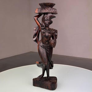 Può includere: Scultura in legno marrone scuro di una donna che porta un cesto sulla testa e tiene un cesto in mano. La scultura è su una base con un cane ai suoi piedi. La scultura è su un tavolo bianco.