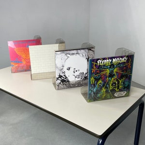 Könnte beinhalten: Vier Schallplatten, die auf einem weißen Tisch mit blauen Beinen ausgestellt sind. Die Schallplatten befinden sich in durchsichtigen Plastikhüllen. Die Schallplattencover zeigen ein pink-orangenes Design, ein weißes Ziegelmauerdesign, ein Schwarzweißdesign und ein buntes Design mit dem Text "FLERRY MELODY".