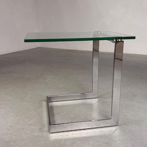 Könnte beinhalten: Ein moderner Beistelltisch mit einer transparenten Glasplatte und einem polierten Chromgestell. Der Tisch hat eine rechteckige Glasoberfläche und ein markantes C-förmiges Gestell. Das Design ist minimalistisch und zeitgemäß.