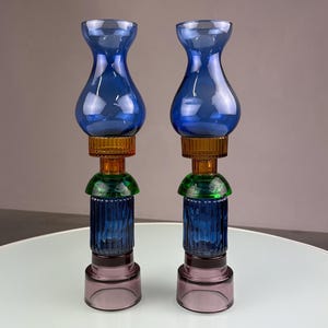 Op de afbeelding: Twee decoratieve glazen kandelaars. Elke houder heeft een blauwe vaasvormige bovenkant, een bruin middengedeelte, een groen accent en een paarse basis. De houders worden op een wit oppervlak weergegeven.