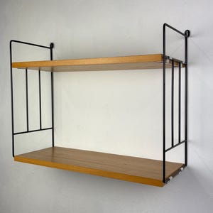 以下が含まれることがあります： 明るい木材の棚板と黒い金属フレームを備えた、2段の壁掛け棚。長方形の棚板は、ミニマリストな黒いフレームで支えられています。白い壁に取り付けられており、アイテムの展示に適しています。